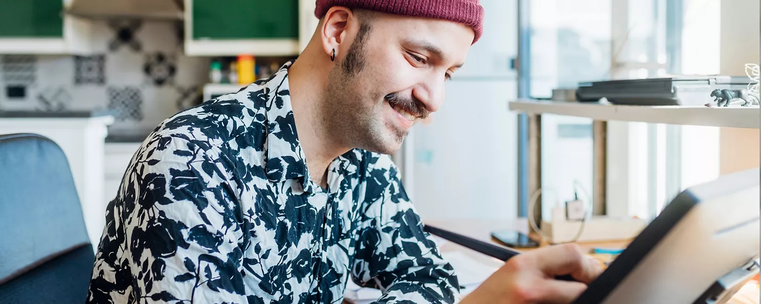 Man wearing a red beanie sitting in a black chair drawing on his computer with a stylus.