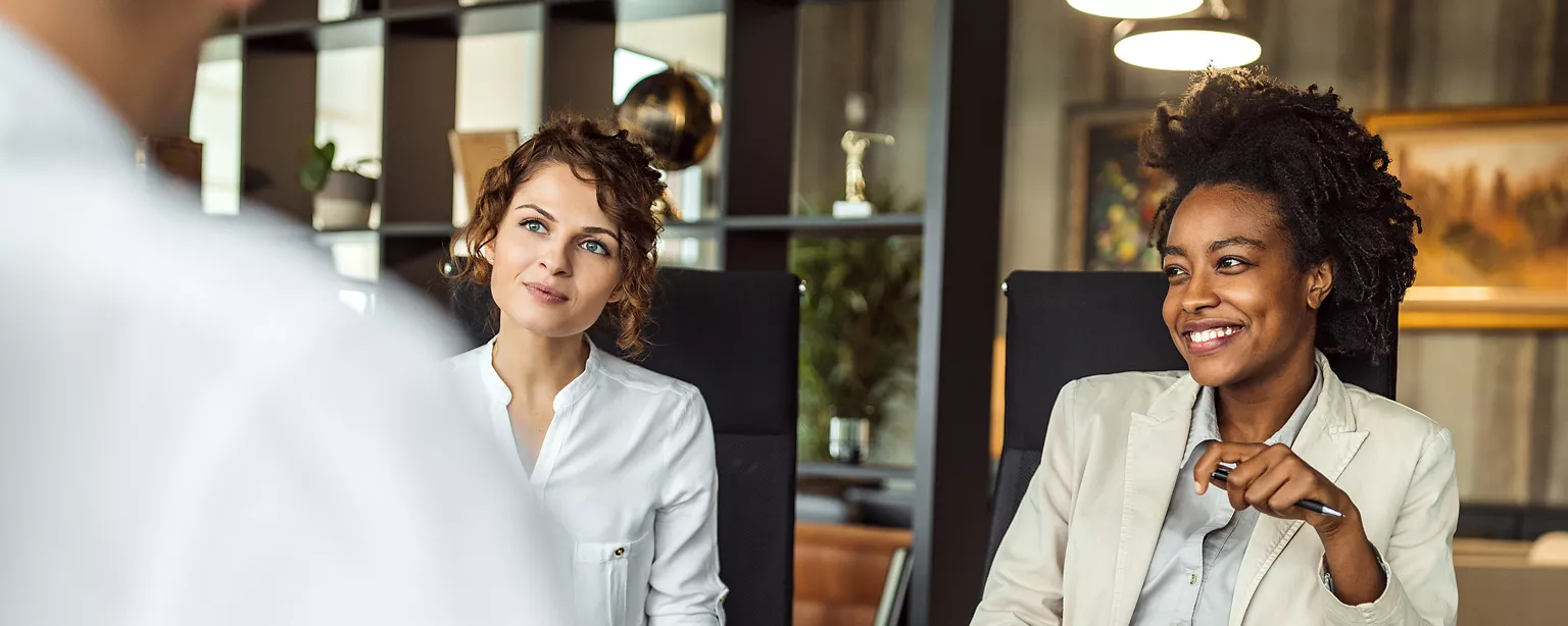 Two professional women are sitting at a table, attentively listening and smiling during a business meeting or interview.