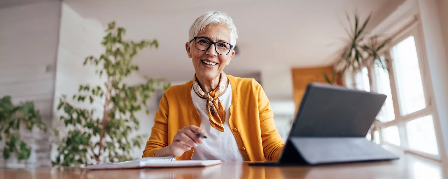 A smiling, mature woman in a yellow sweater sits at a desk in front of a laptop in an office.