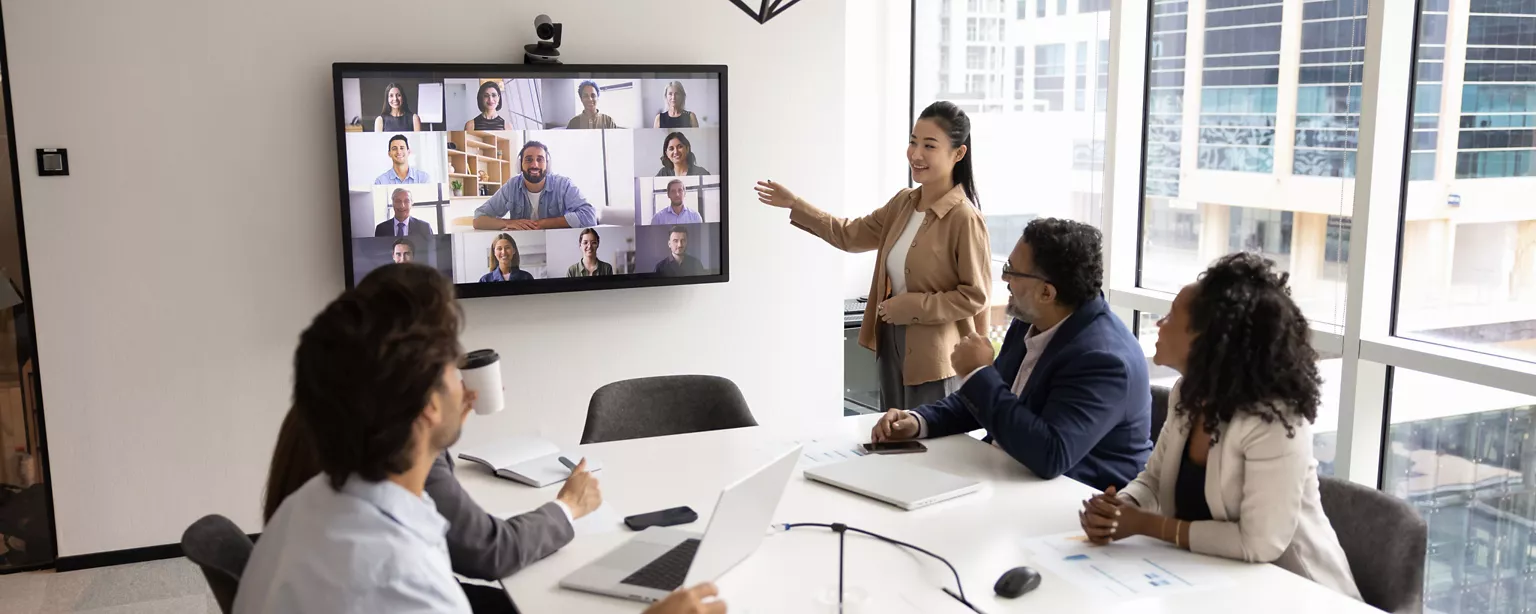 Team meeting in a modern office with in-person employees watching a large screen displaying remote colleagues on a video call.