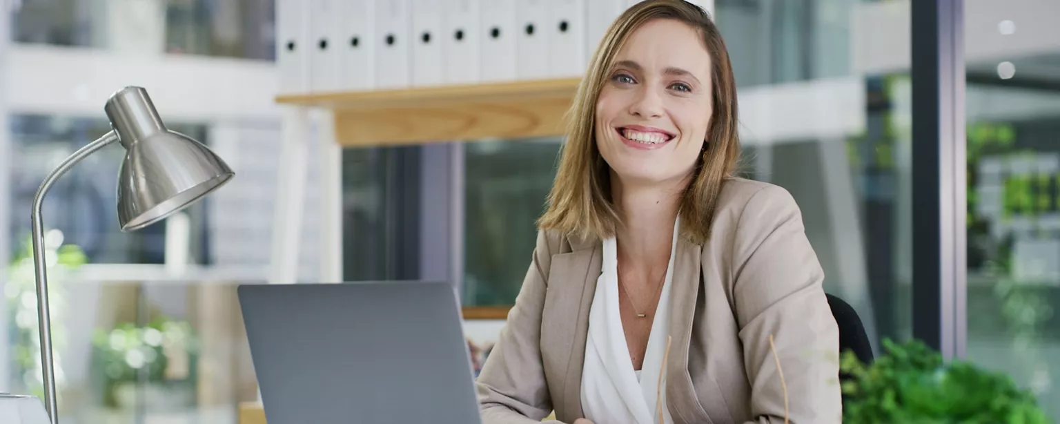 A businesswoman looks at us while smiling and sitting at her desk with an open laptop.