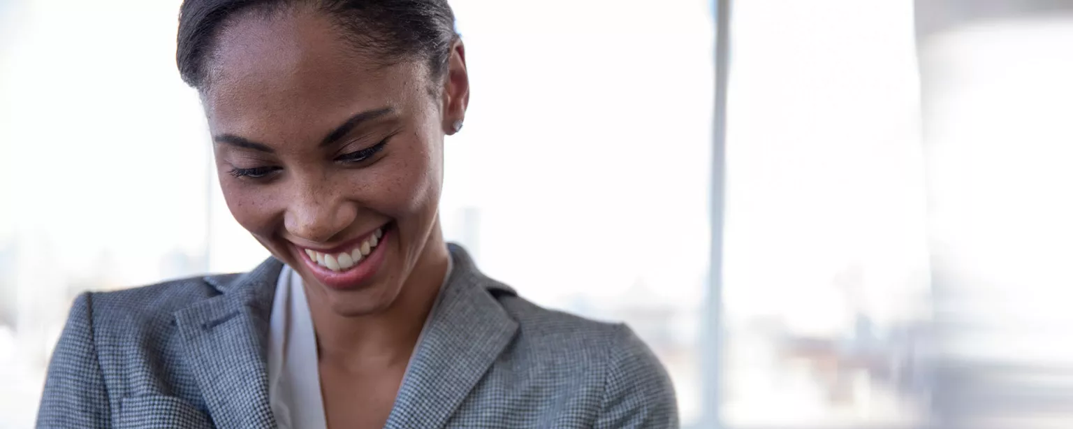 A businesswoman in a grey blazer and dress, standing in front of a conference room, tapping the screen on her smartphone and smiling.