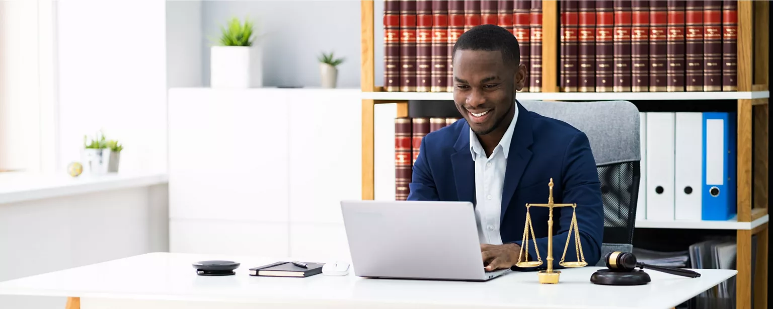 A smiling legal professional in a suit working on a laptop at a desk with law books, scales of justice and a gavel.