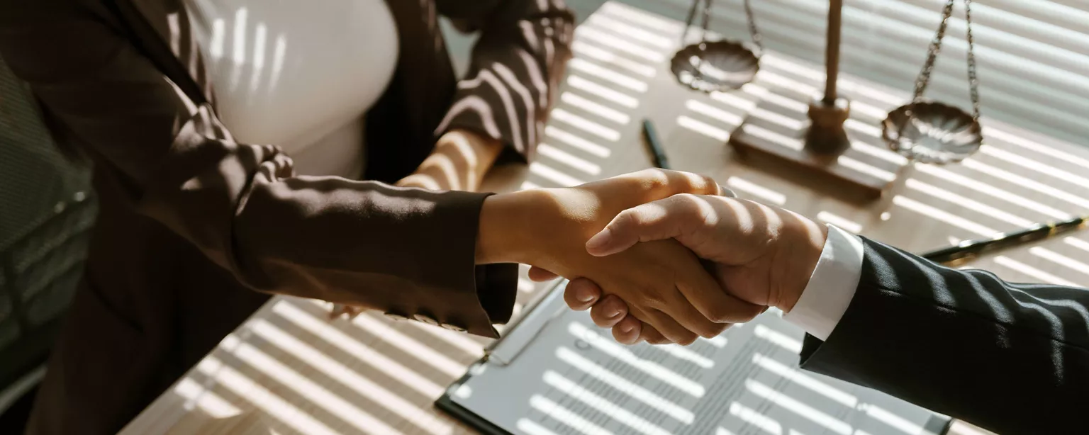 Two business professionals shake hands across a desk with legal documents and scales of justice in the background.