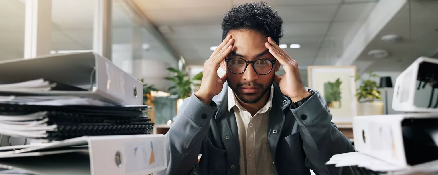 A finance professional sits at a desk with stacks of binders, holding his head in stress.