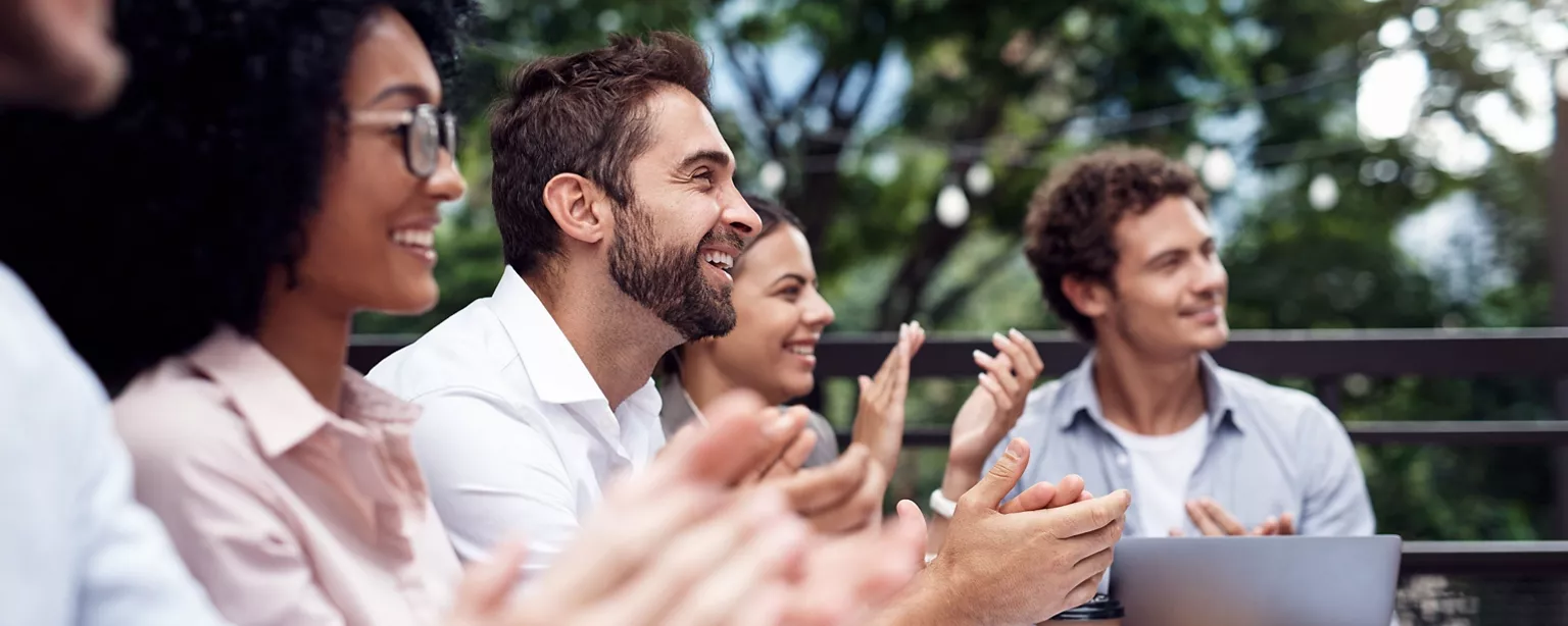 A diverse group of colleagues seated outdoors is smiling and applauding during a team meeting or celebration.