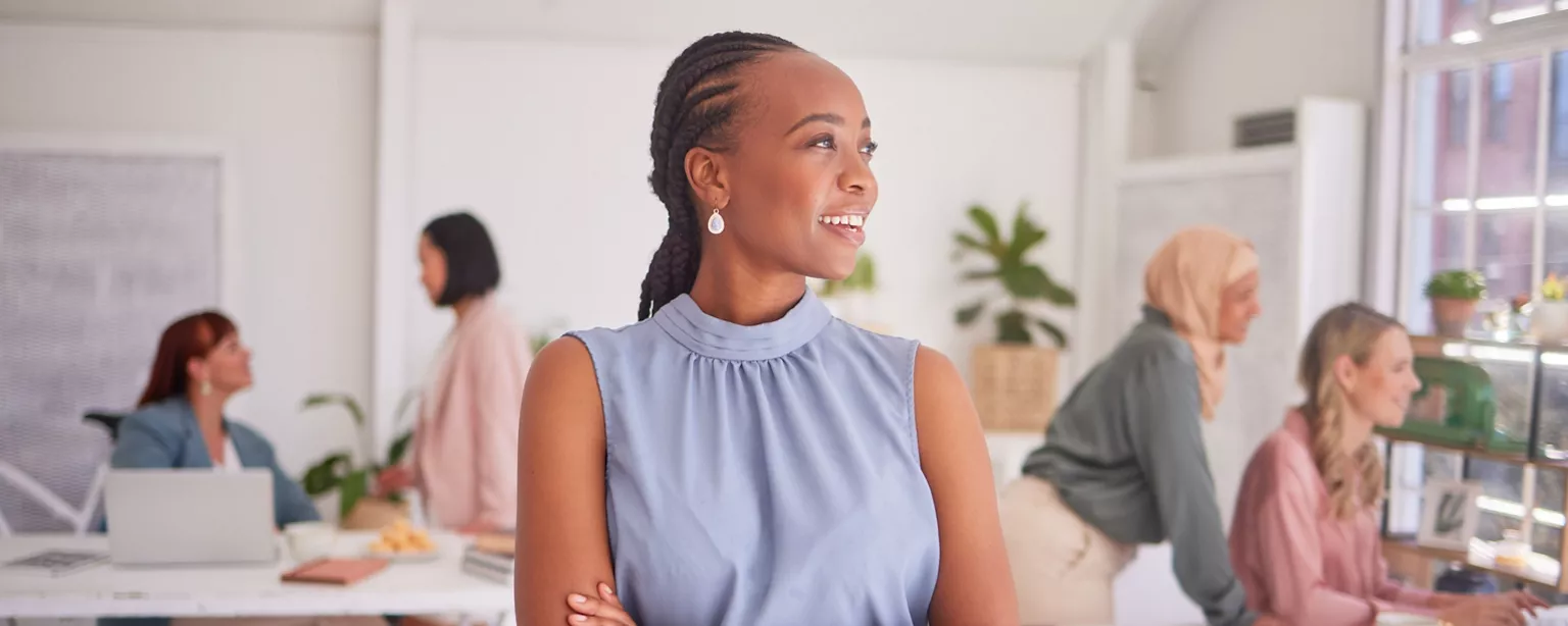 A confident woman smiles and looks out the window in a bright office space while colleagues collaborate in the background.