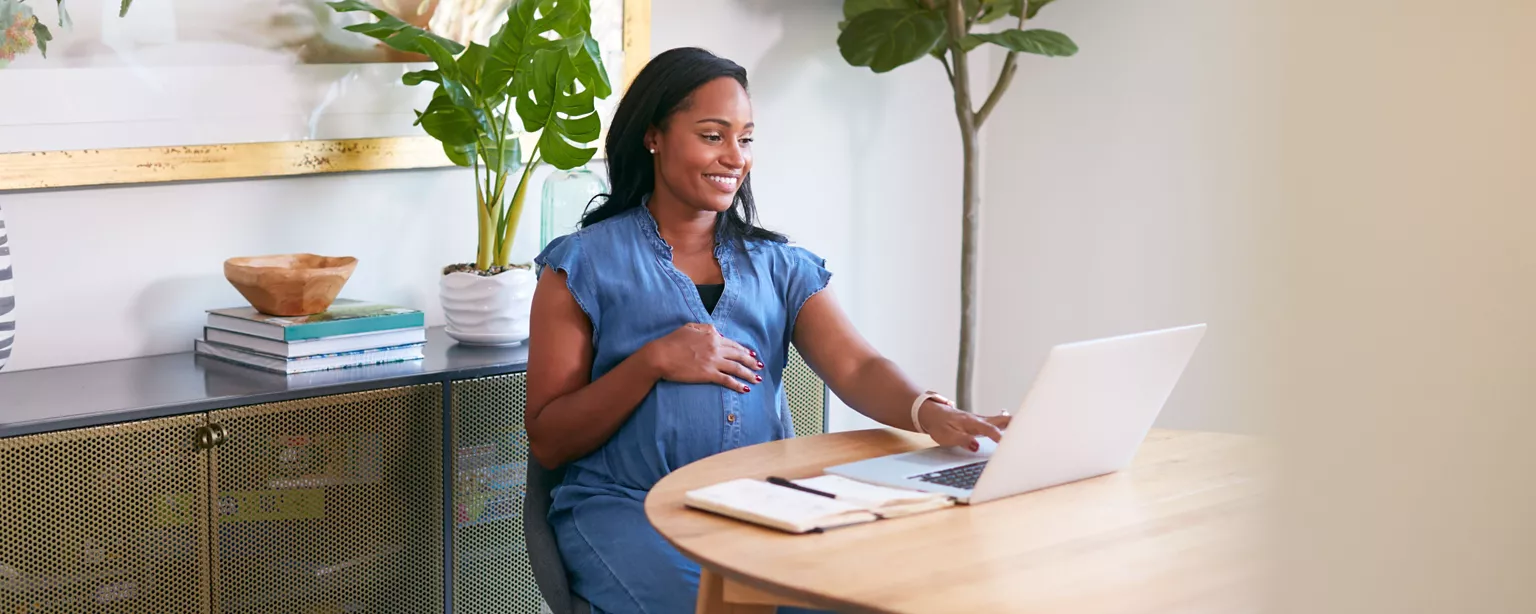 A pregnant woman, seated at a table, smiling as she works on a laptop, with a notebook nearby and plants in the background.