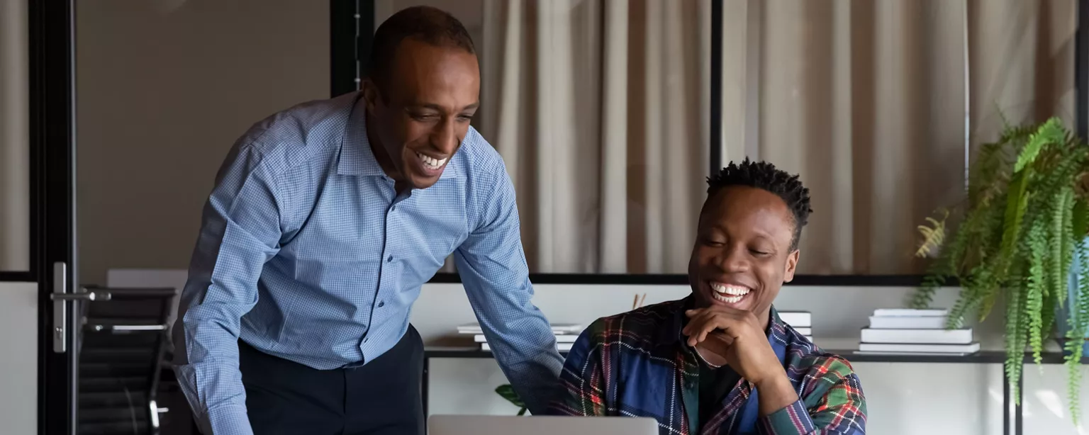 Two colleagues smiling and collaborating in an office setting, with one providing guidance to the other while looking at a laptop.