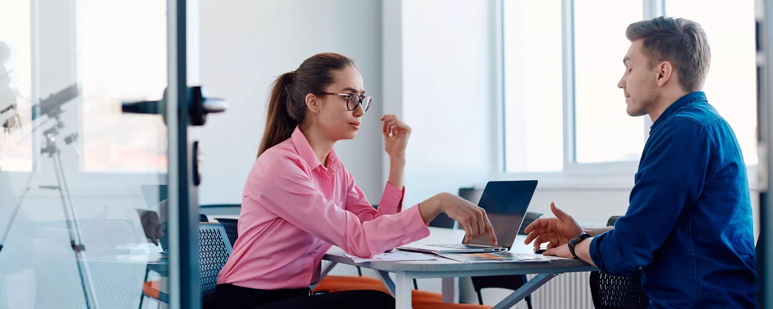 Two business professionals, a woman in a pink shirt and glasses and a man in a blue shirt, are having a focused discussion at a table in a conference room.