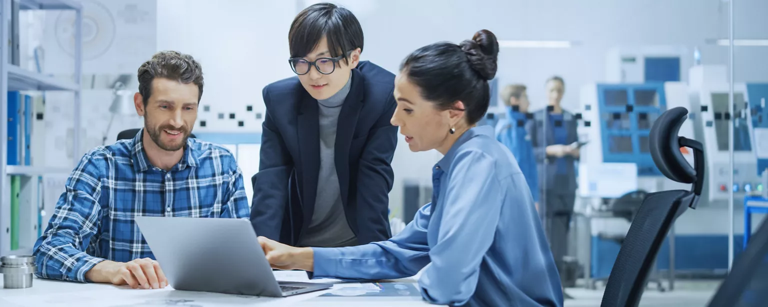 Three professionals in an office collaborate over a laptop, discussing project details and reviewing documents together.
