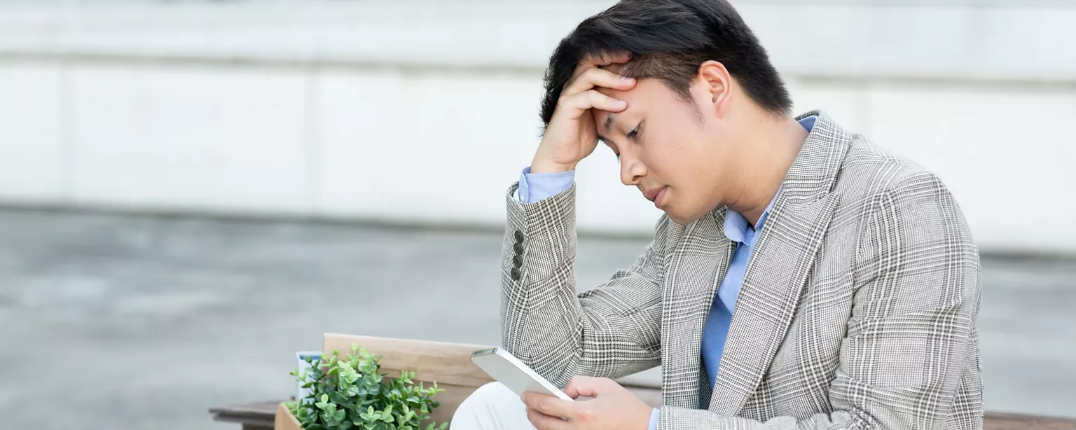 A man stares at his phone as he sits dejectedly on a bench in an urban environment, head in hand, demotivated by being ghosted after a job interview.