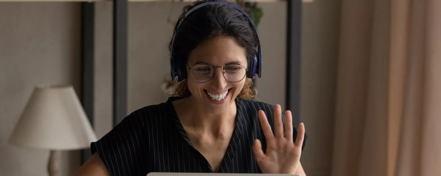 A smiling woman waves at her laptop during a video call.