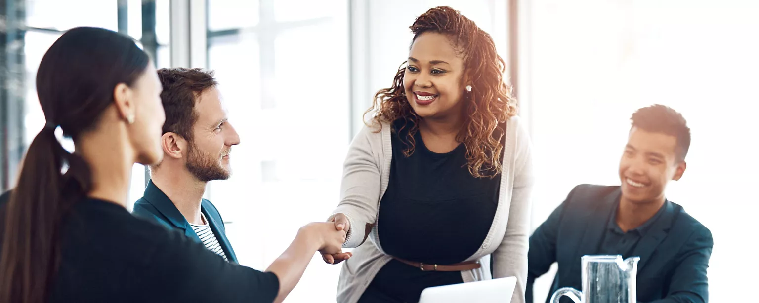 A group of colleagues smile and connect during a team meeting, with one woman warmly greeting a coworker with a handshake.
