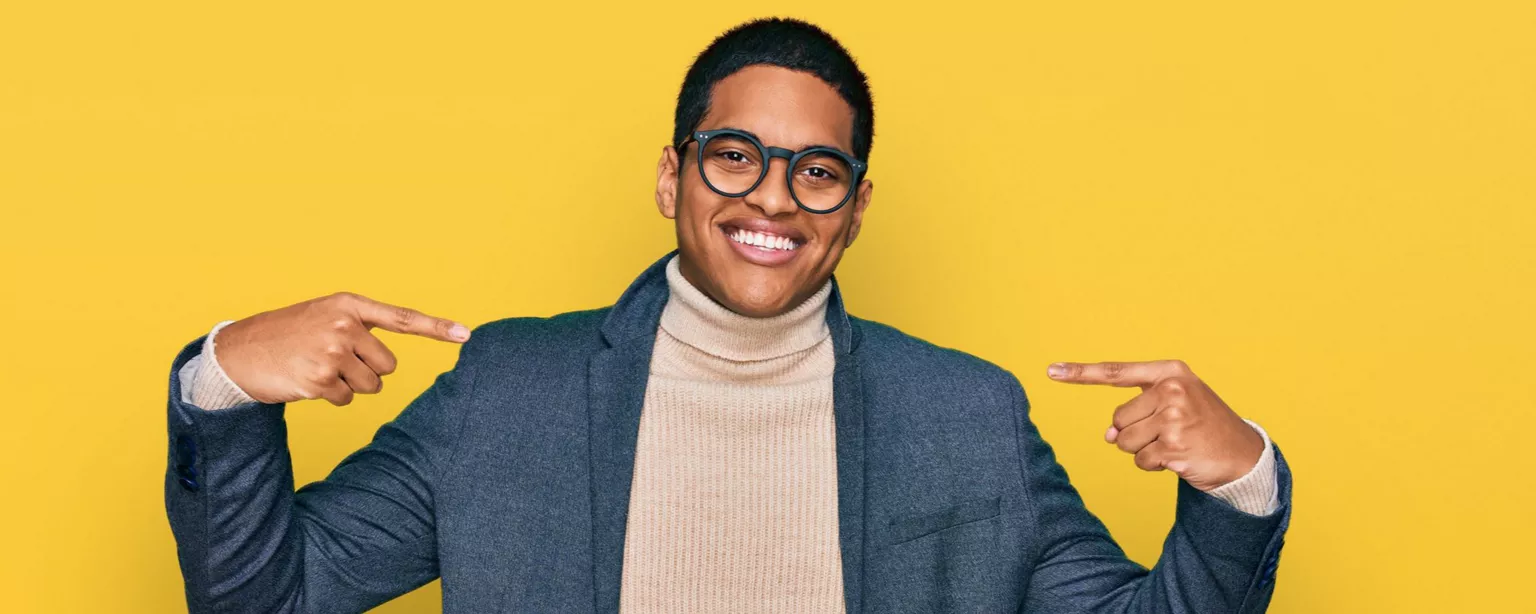 Smiling young man wearing glasses, a turtleneck and a blazer, pointing to himself confidently with both hands.