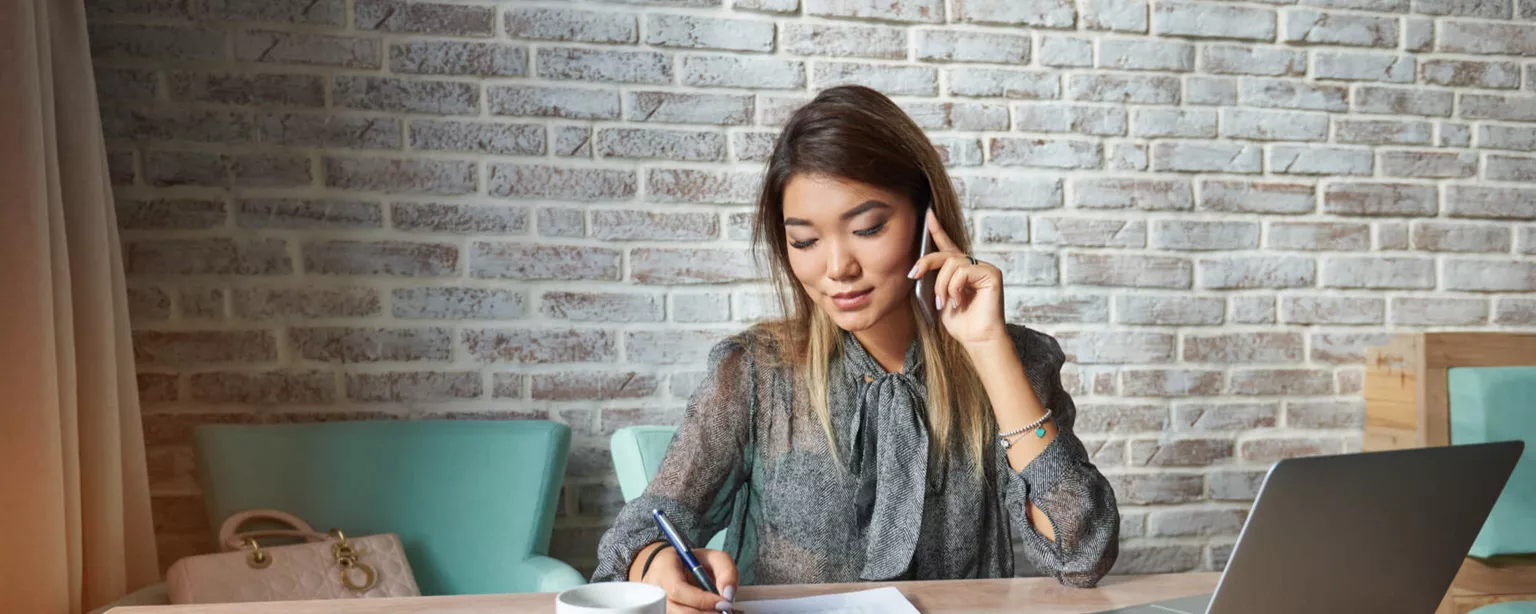 A woman takes notes as she listens with her ear to the phone at a table in an office.