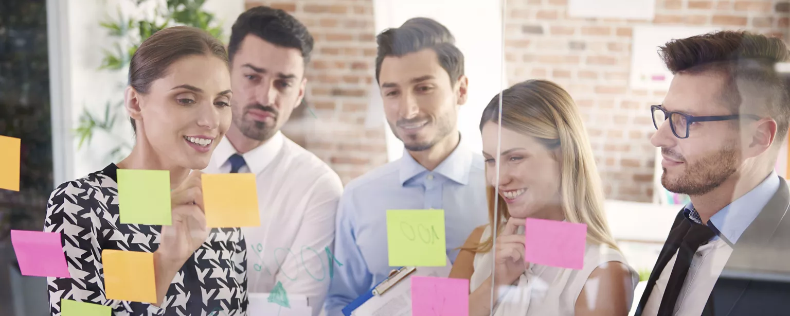 A group of professionals collaborates in front of a glass wall covered with colorful sticky notes, brainstorming ideas in a bright office.