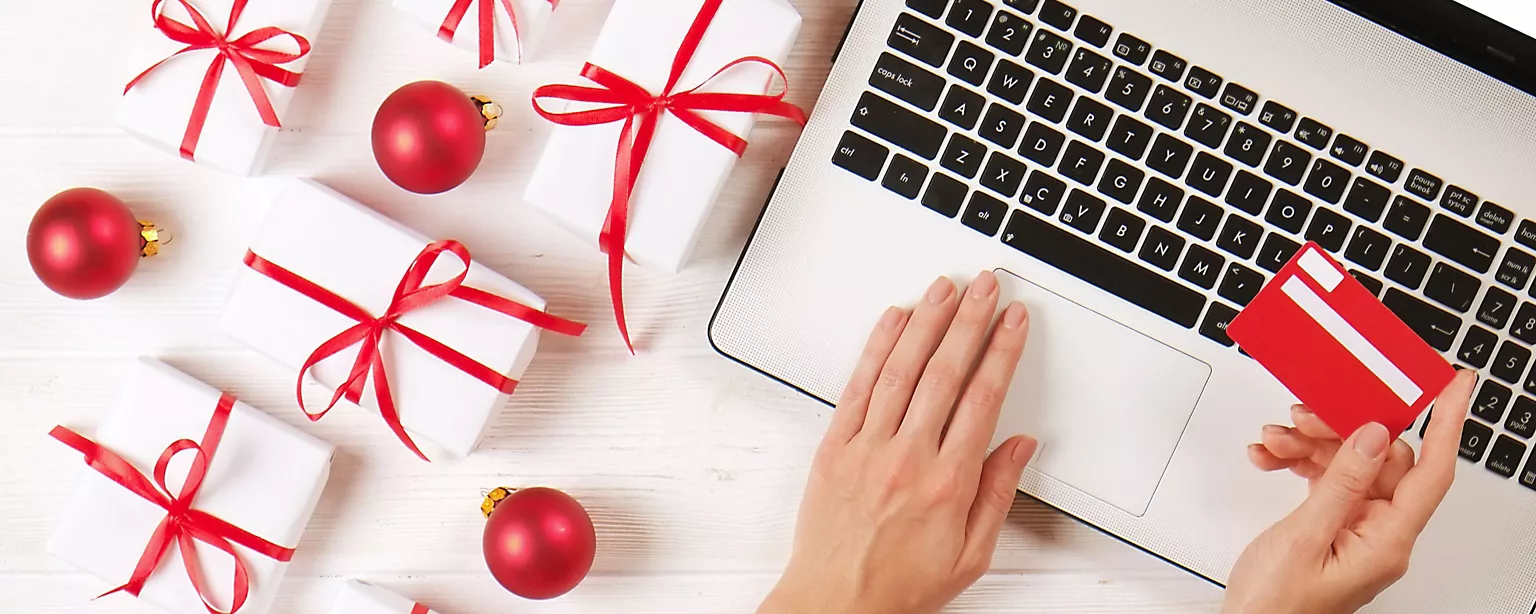 Overhead view of hands typing on a laptop keyboard while shopping online with a credit card, surrounded by holiday gifts wrapped in white paper with red ribbons and red ornaments.