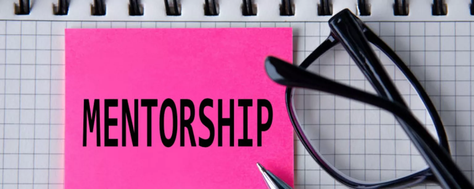 The word 'mentorship' appears on a pink sticky note next to eyeglasses and a pen on top of a notebook.
