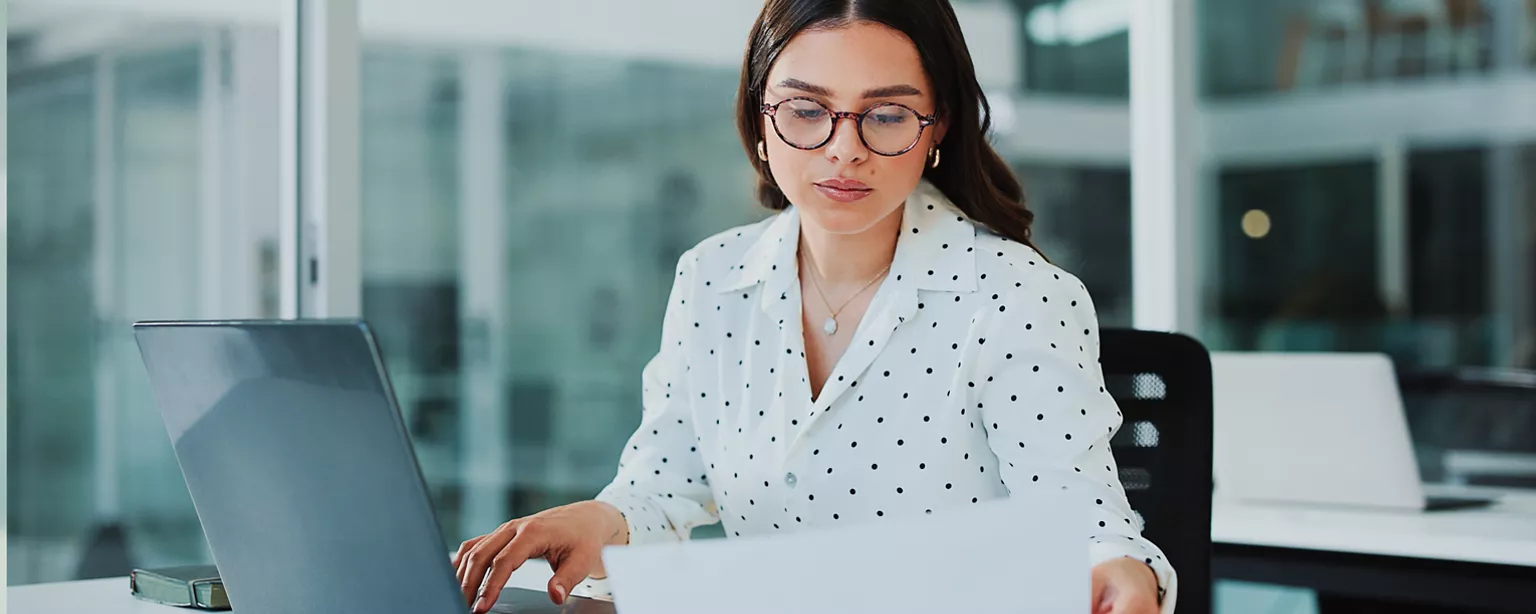 a woman at work in a modern office, working on her laptop whilst glancing at a piece of paper to her left