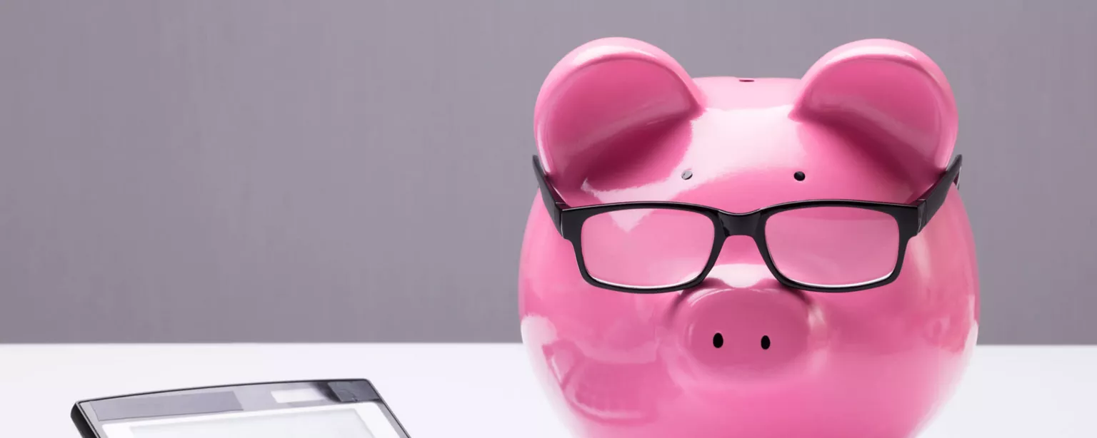 A calculator on a table, next to a pink piggy bank with black-framed glasses balanced on the pig's snout.