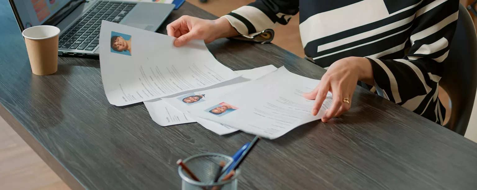 A hiring manager's hands are seen holding resumes featuring photos of three diverse job candidates.