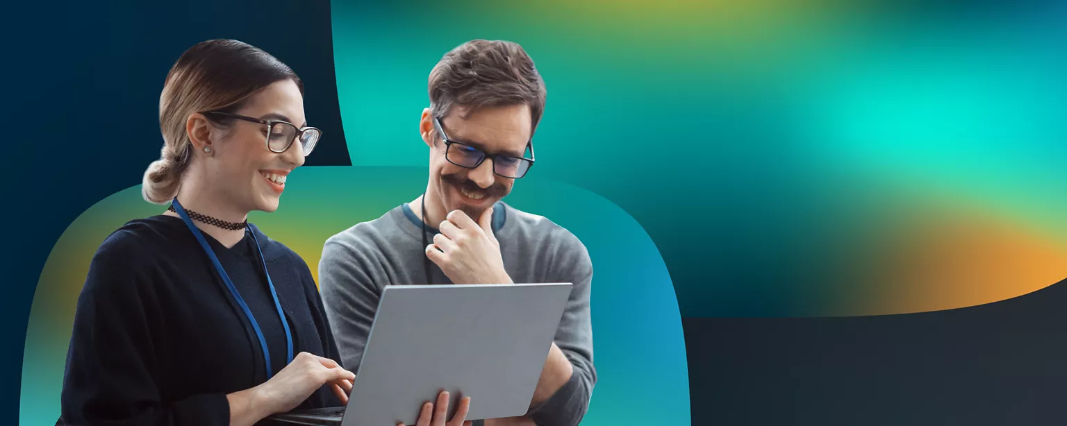 A woman and a man wearing glasses stand next to each other smiling and looking at a laptop.