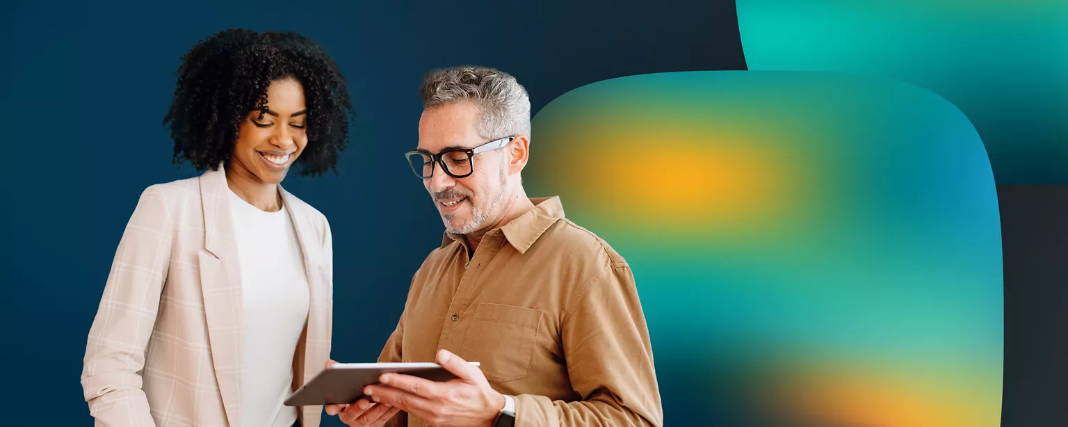 A woman with black curly hair wearing a cream-colored blazer stands next to a man in glasses wearing a brown button-down collared shirt while looking at a tablet.