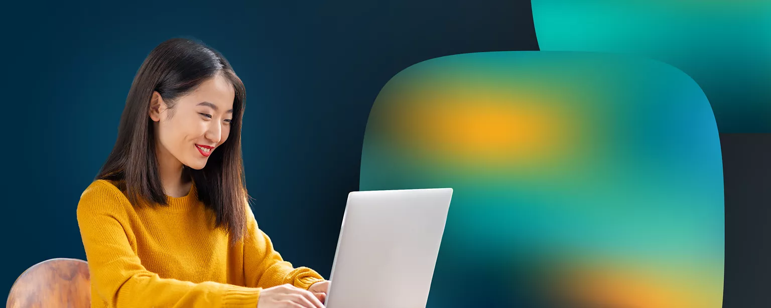 A woman in a yellow sweater sits at a desk smiling while looking at a laptop.
