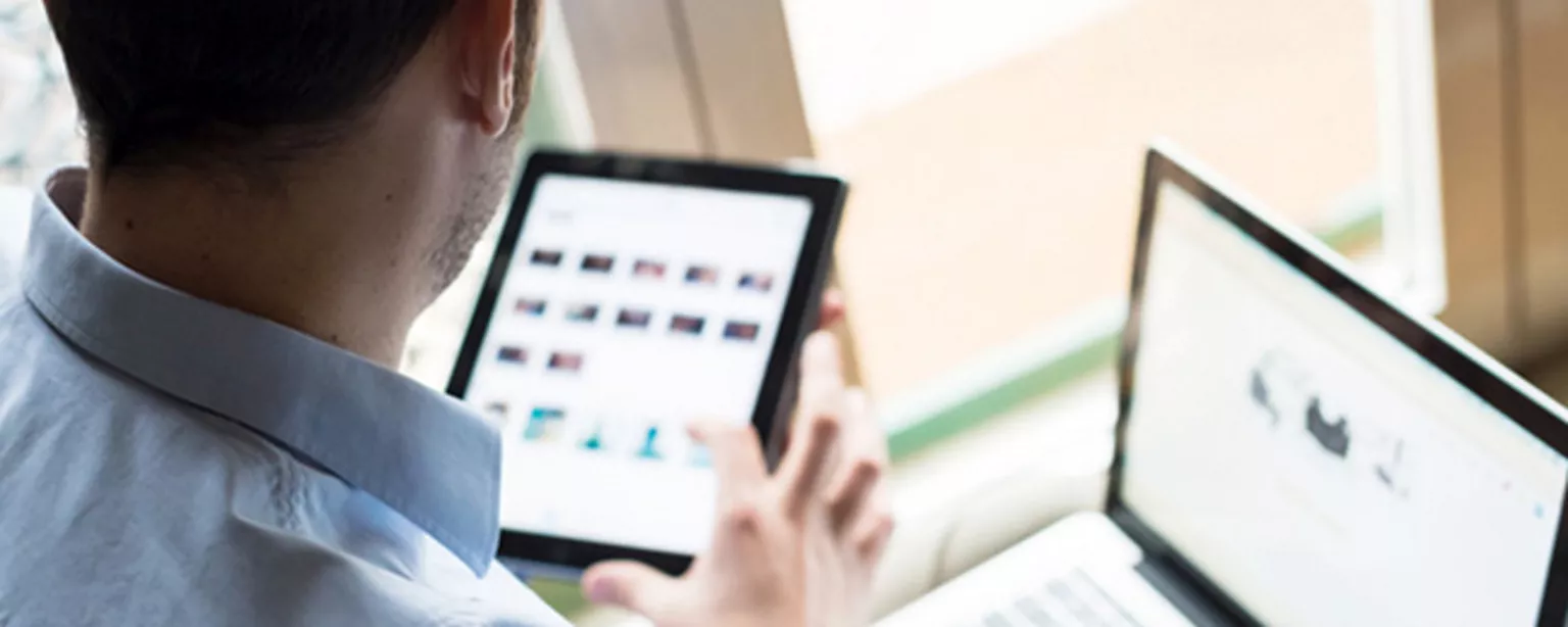 Man holding a mobile device with a laptop computer in the background.