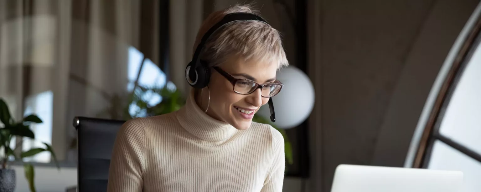 Woman with short hair wearing glasses and a headset, holding a pen and smiling while looking at her laptop screen while working remotely.