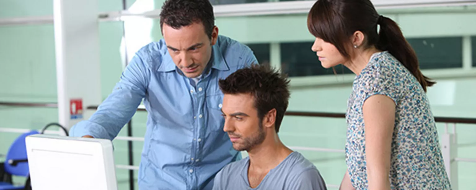 A man and a woman stand on either side of a man seated in front of computer, all of them looking at the monitor. 