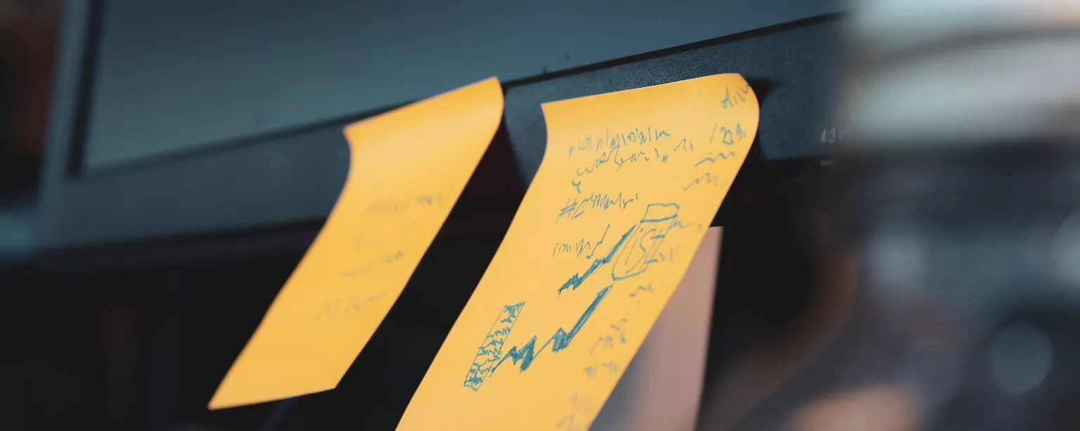 Gelbe Haftnotizzettel mit schwer entzifferbaren Notizen kleben am Rand eines Computerbildschirms