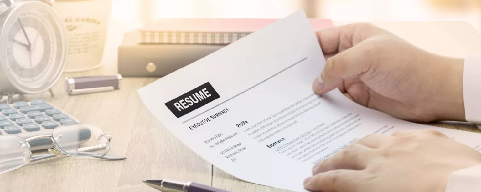 Hands hold a printed resume next to pen on a desk.
