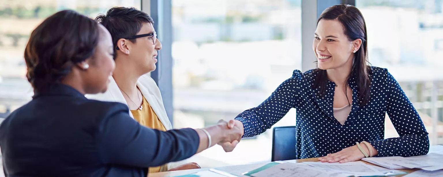 Three professionals sit together at a meeting table with two of them shaking hands and smiling.