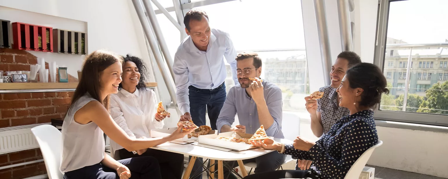 A small business team enjoys a casual lunch, smiling and connecting over a team pizza lunch in a bright office.