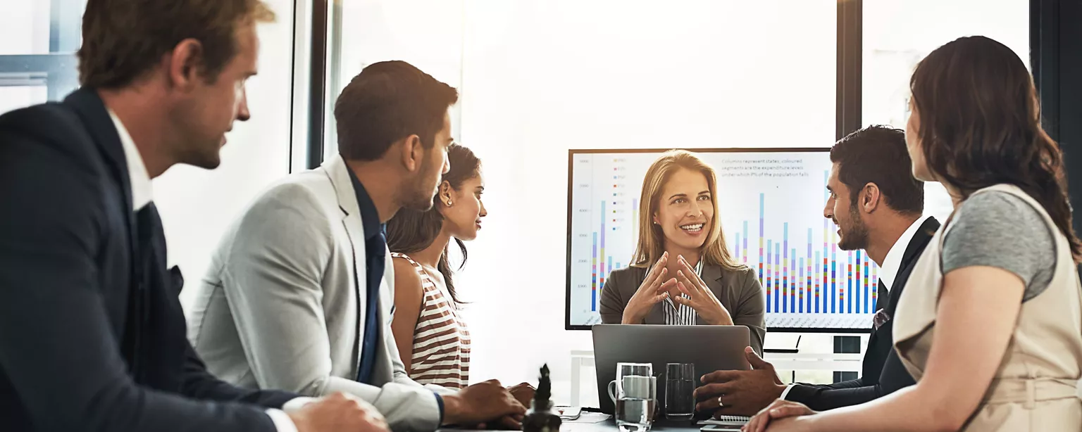 Business professionals in a meeting room engage in a strategic discussion, with a woman leading the conversation in front of a screen displaying data charts.