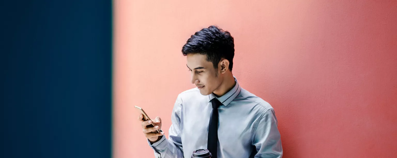 Mann lehnt mit einem Kaffee in der Hand an eine Wand und sieht auf sein Handy