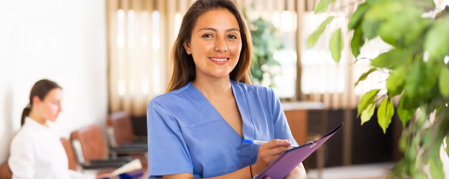 A healthcare professional in blue scrubs smiles while holding a clipboard in a bright, welcoming office environment.