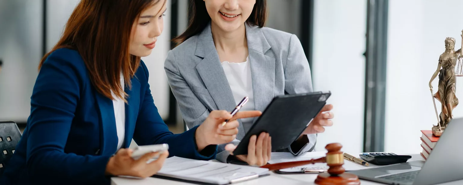 Two young lawyers sit at a desk with a gavel, a statue of Lady justice, books and a laptop, discussing documents on a tablet.