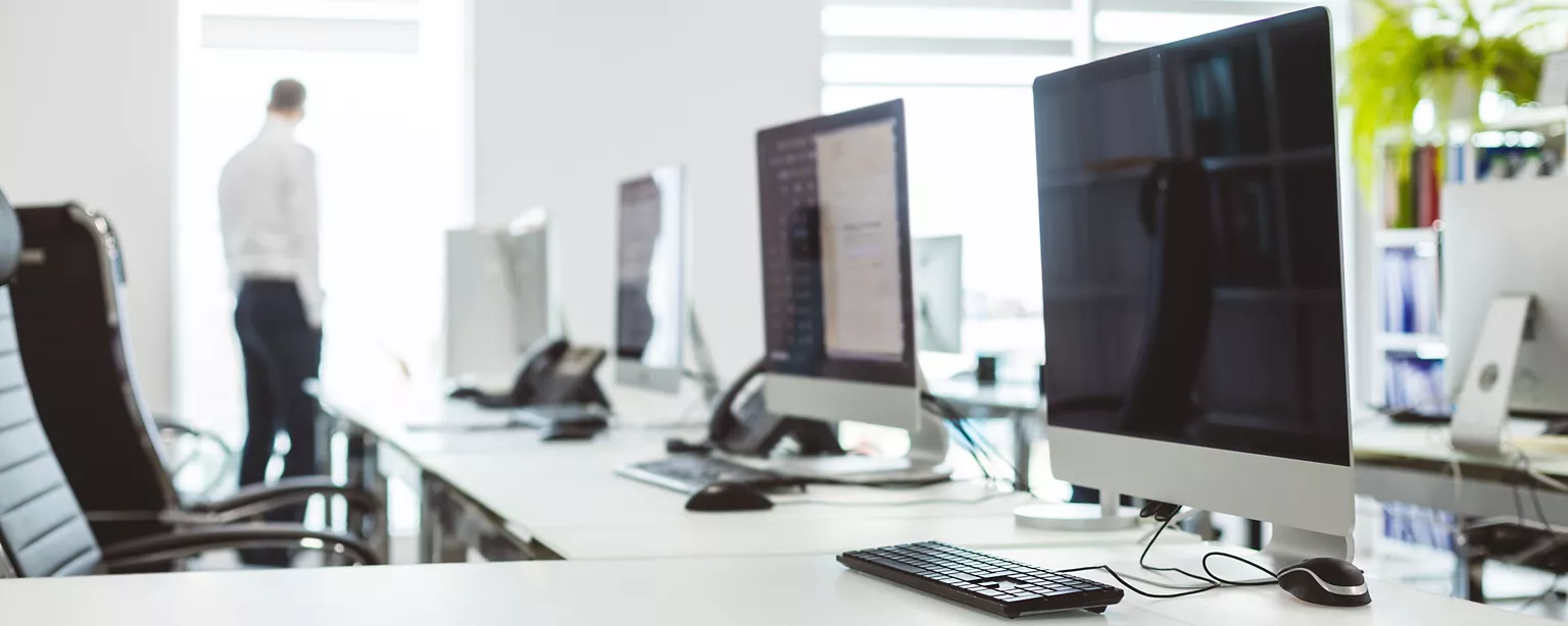 An empty office workspace with multiple desktop computers and office chairs, with a person standing by a window in the background.