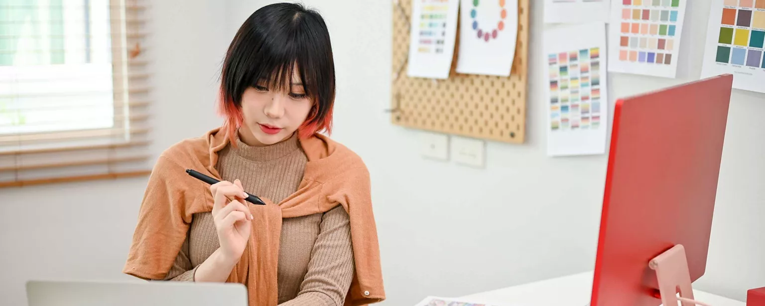 A graphic designer works at a desk with color swatches pinned on the wall behind her and focuses on a digital pen tablet in front of her.