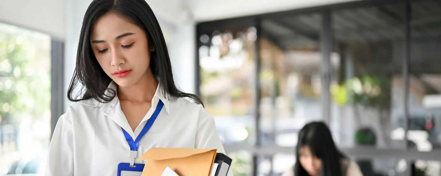 A young woman with a sad expression on her face looks downward as she carries a box of her belongings out of the building.