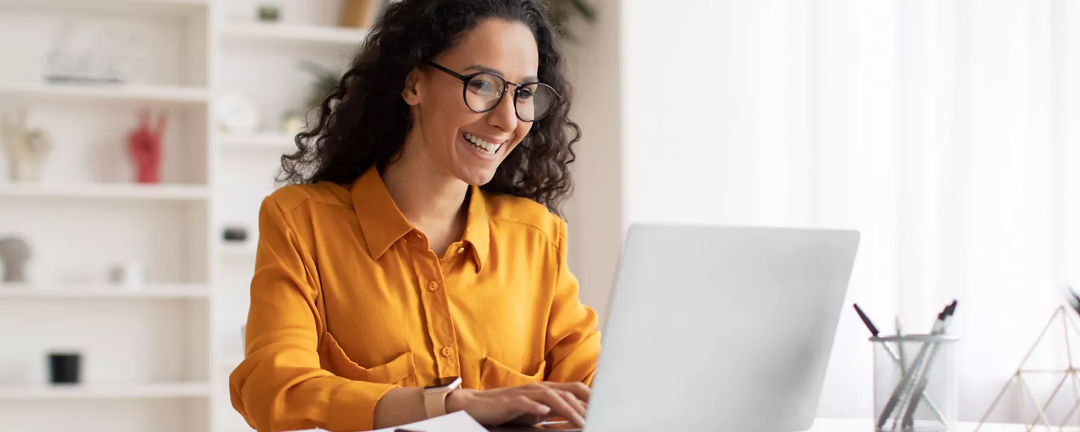 A smiling woman in a mustard-colored blouse works on her cover letter on her laptop in a bright home office.