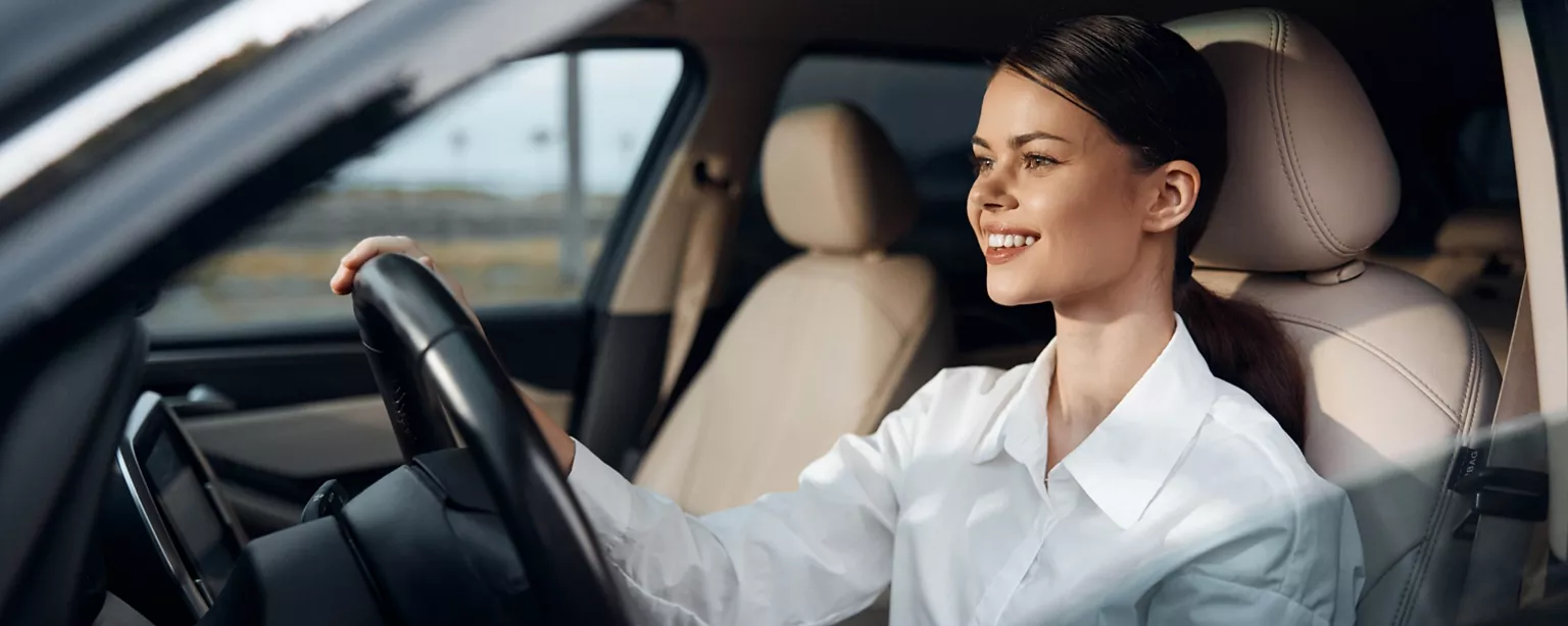 A woman in a white button-up smiles confidently while driving a car on her way to work.