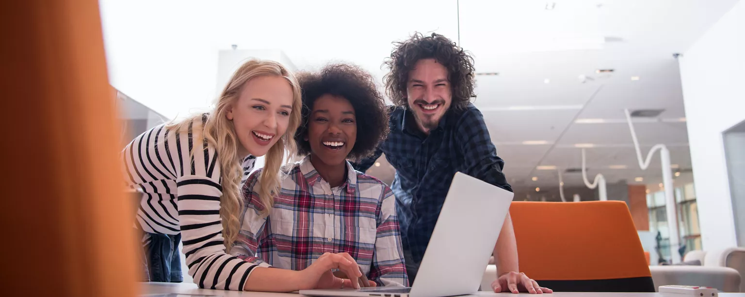 Three diverse individuals smile at work, with one person using the keypad on a laptop to input information while the other two watch her type. 