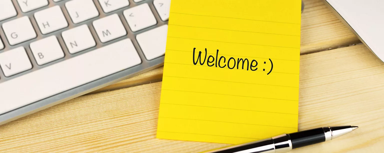 The word "Welcome" appears on a yellow sticky note next to a computer keyboard and a pen on a desk.