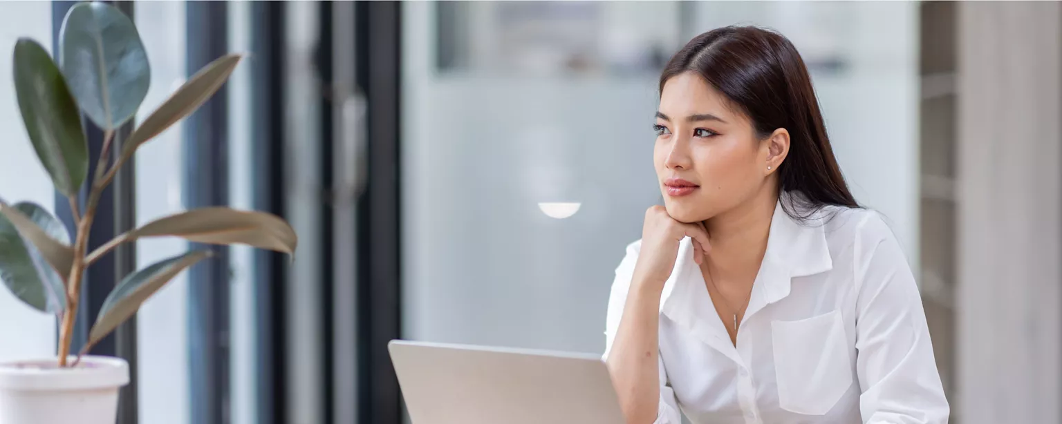 A professional woman in a white blouse sits at a desk, thoughtfully looking off into the distance with a laptop in front of her.