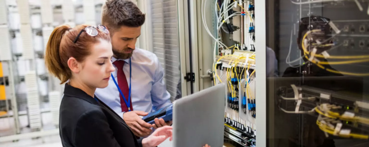 Two systems engineers use their laptops while analyzing a server.