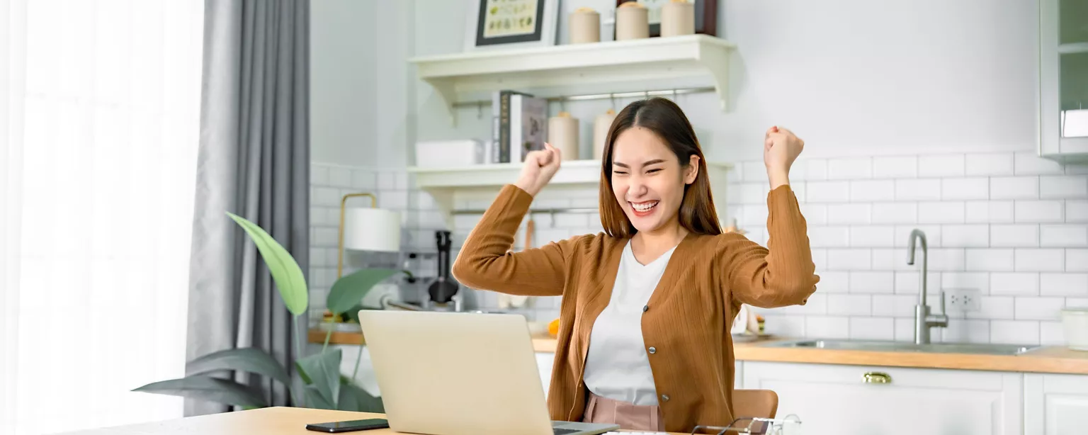 A woman smiles and raises her arms in celebration while looking at a her job offer on a laptop in a bright kitchen.